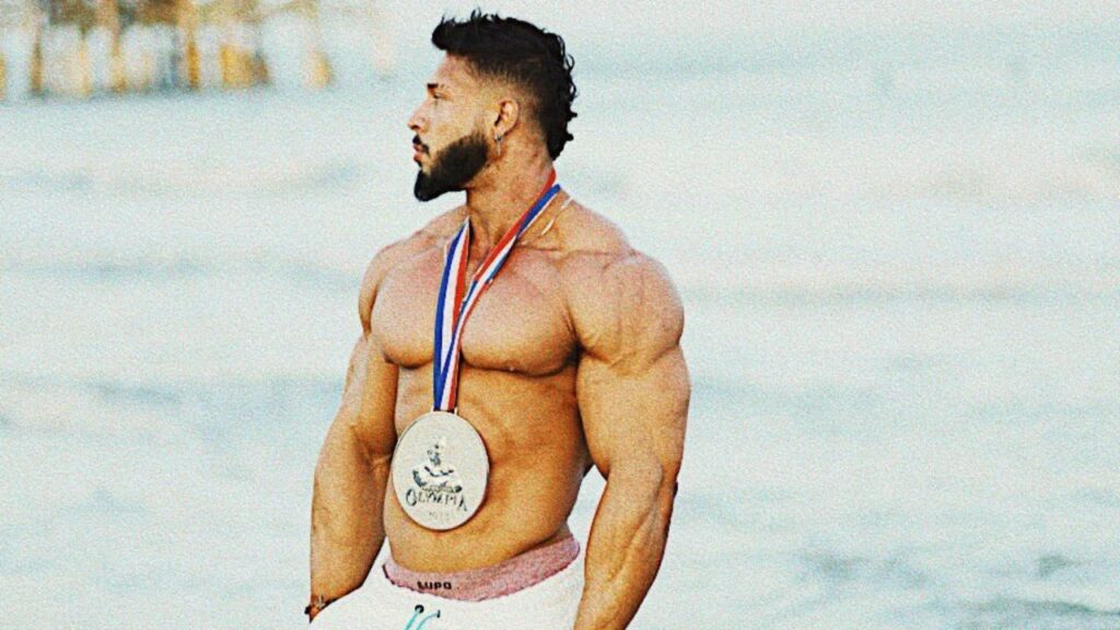 Atleta da Max Titanium, Ramon Dino, olhando para o horizonte na praia e carregando a medalha do título do Mr. Olympia em seu pescoço